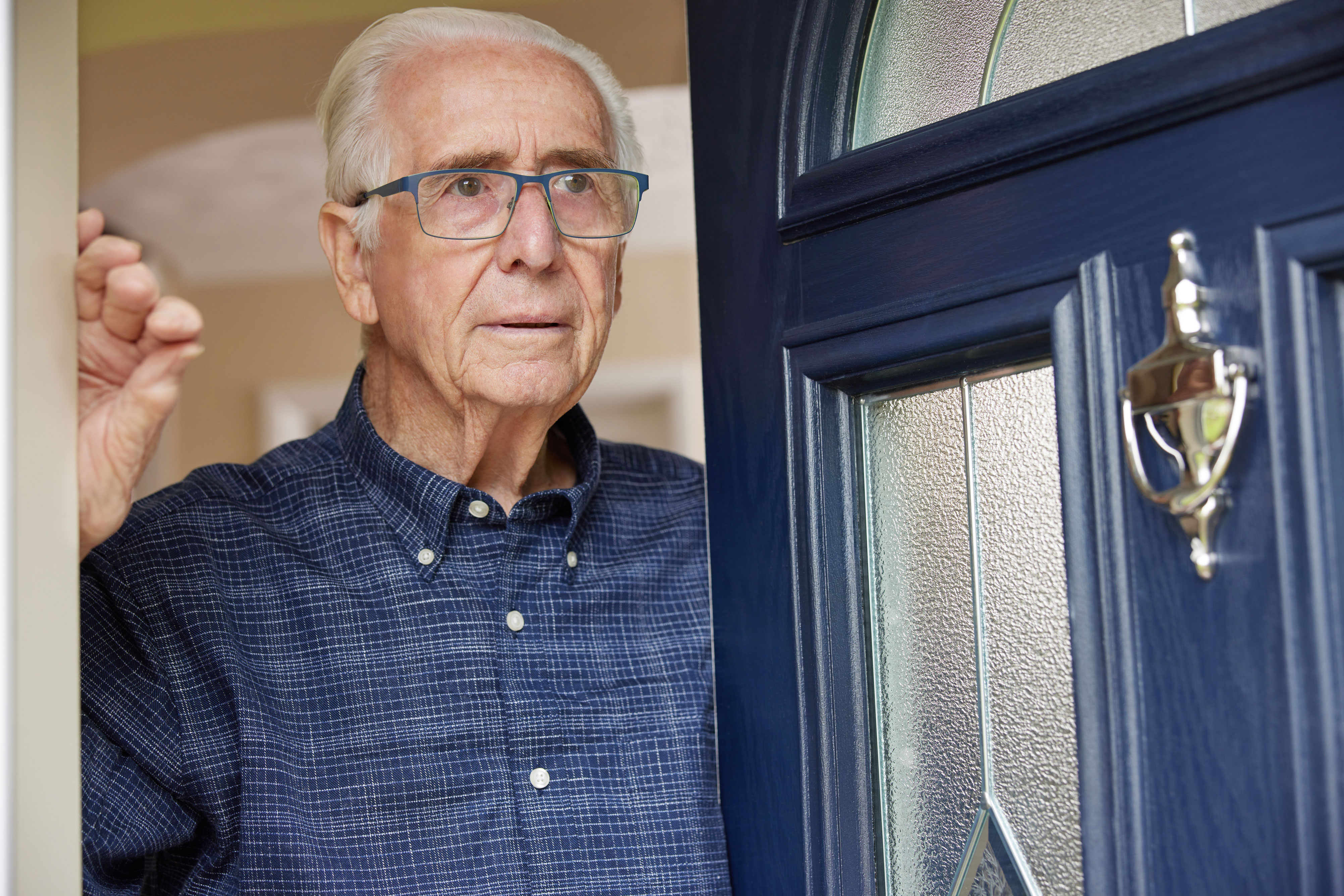 Old man with a blue checked shirt and blue glasses opens the blue front door and looks outside. The front door has window inserts and a door knocker.