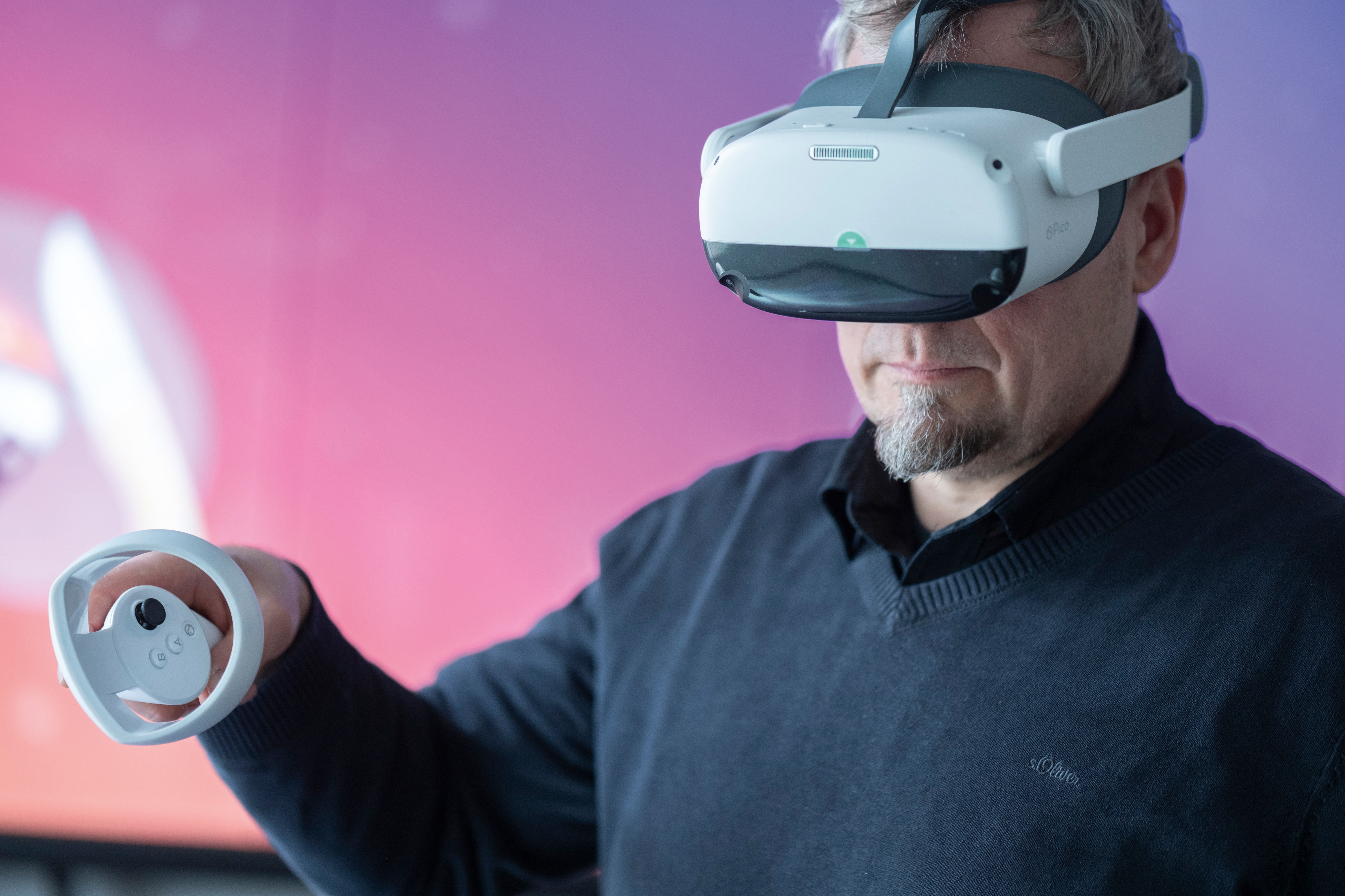 A man wears VR goggles and holds a controller in his hand. A screen with a pink color gradient can be seen in the background.