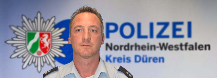 Bezirksdienstbeamter Oliver Pfeiler vor Leinwand mit dem Logo der Polizei Düren