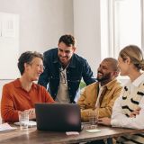 Ein freundliches Büro mit Holztisch. Vier Kollegen, zwei Männer und zwei Frauen, unterhalten sich angeregt und lachen.