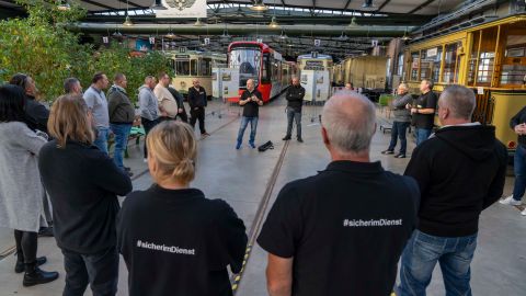 You can see a hall in which streetcars are stored. In the foreground is a group of people in a circle. Two people are wearing T-shirts with the inscription #Safe on duty. One person is explaining something to the group.